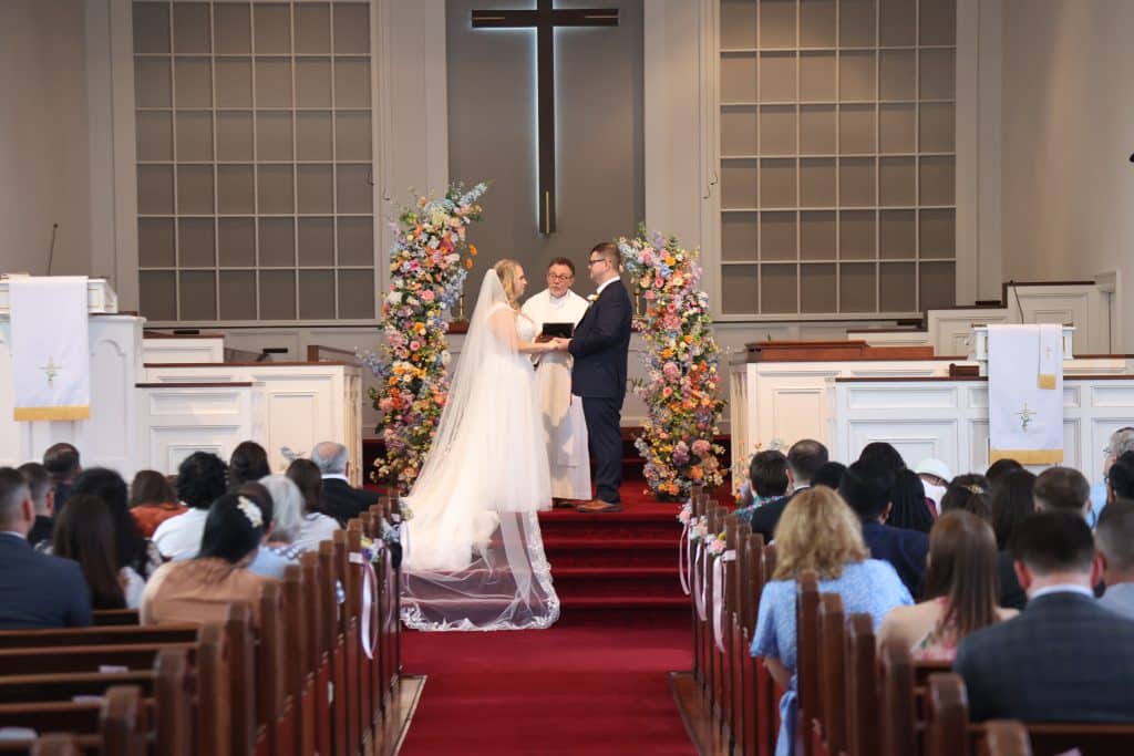 Leila and Derrick standing at the altar in front of vibrant floral arrangements during their wedding ceremony.