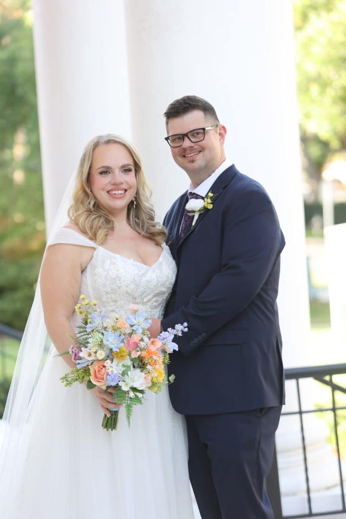 Leila and Derrick smiling together outdoors with a colorful wedding bouquet.